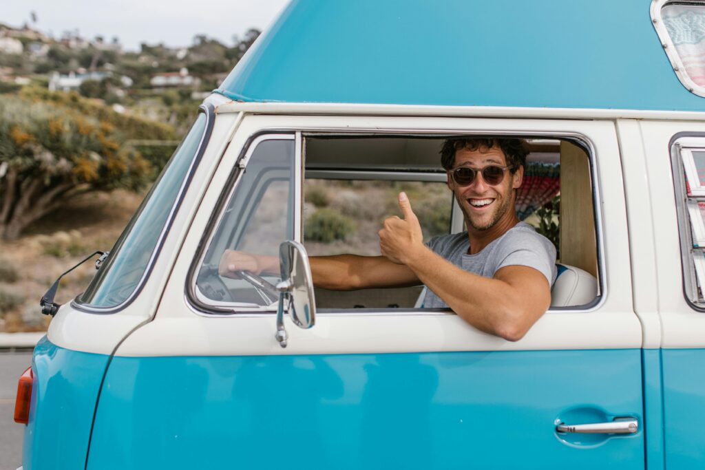 Smiling man giving thumbs up while driving a vintage blue camper van outdoors.